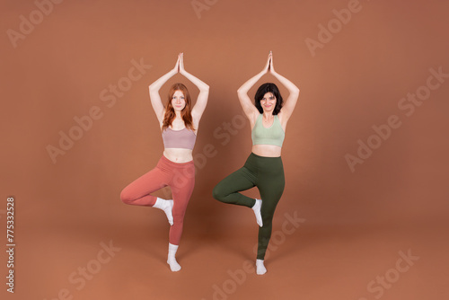 Two young women practice yoga in the Tree Pose, dressed in sportswear, showcasing balance and serenity against a brown backdrop.