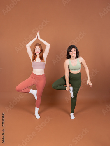One young woman gracefully holds the Tree Pose while the other smiles and attempts to mirror the pose, showcasing determination and camaraderie in their yoga practice.
