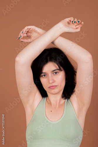 A young woman with black hair, wearing a green sports bra, crosses her arms over her head and looks directly at the camera against a brown background, exuding confidence and strength.