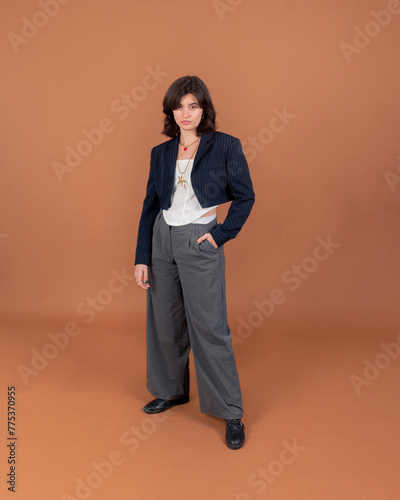 A young brunette woman dressed in a casual style with a blazer and dress pants against a brown backdrop, exuding an air of sophistication blended with comfort.