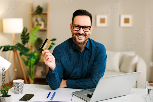 Young happy man is holding a credit card in his hand while sitting at a table with a laptop in front