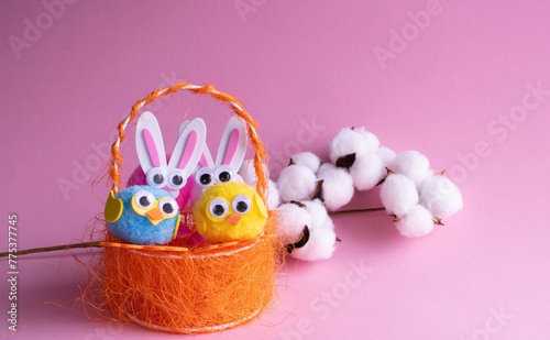 A branch of white cotton, toy eggs with birds in an orange basket and Easter bunnies on a pink background.