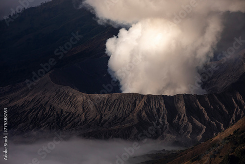 An imposing volcano belches a cloud of steam and ash into the sky, standing stark against a backdrop of soft morning mist photo