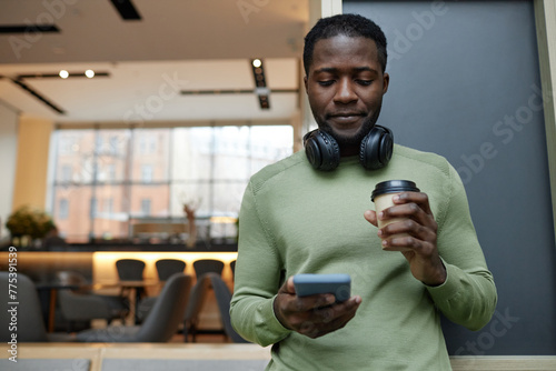 Minimal waist up portrait of young African American man using smartphone leaning on wall in office lounge and drinking coffee copy space