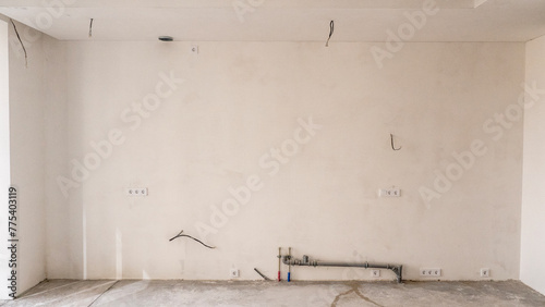 The room is under construction with a rough finish, plastered walls, concrete floor. Wiring of ventilation pipes on a concrete ceiling in a house under construction