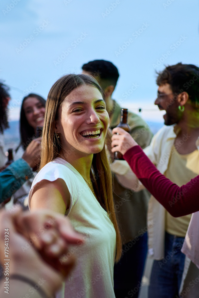 Naklejka premium Vertical. POV young smiling Caucasian woman holding hands boyfriend at party. Attractive girl looking cheerful on camera friends in background outdoors. People gathered on rooftop celebrating birthday