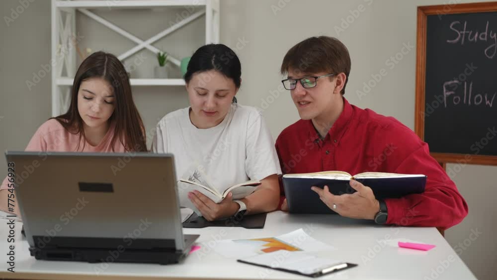 A guy and two girls look at their lecture notebooks and compare them with the data on the laptop screen on the table. Young people are preparing for an exam, sitting at a table in the living room A guy and two girls look at their lecture notebooks and compare them with the data on the laptop screen on the table. Young people are preparing for an exam, sitting at a table in the living room