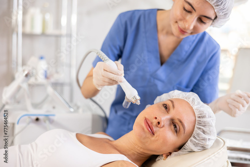 Young woman cosmetologist performs facial moisturizing procedure with machine to young female client