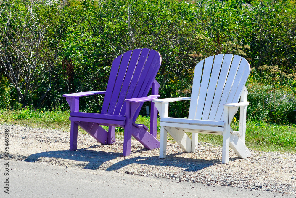Vibrant empty purple and white color Adirondack chairs in a lush green grassy garden. The chairs have curved backs with slats and are on the side of a road with green shrubs behind the chairs. Vibrant empty purple and white color Adirondack chairs in a lush green grassy garden. The chairs have curved backs with slats and are on the side of a road with green shrubs behind the chairs.