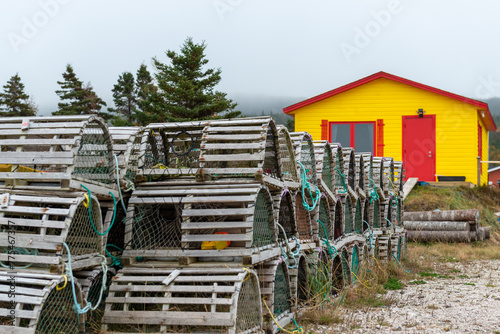 Rows of traditional wooden lobster pots and traps arestacked on a grassy meadow. The cages are made of small wood sticks, green nylon fishing rope. There's a yellow storage building with a red door.   photo