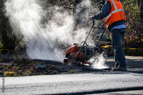 Construction worker using a plate compactor to finish the fresh asphalt at the end of a sidewalk while early morning sun highlights the steam and smoke, road re-paving project 