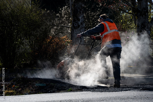 Construction worker using a plate compactor to finish the fresh asphalt at the end of a sidewalk while early morning sun highlights the steam and smoke, road re-paving project
