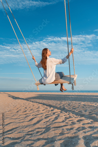 Blond young woman with long hair and white shirt sits on swing above sea. Magic sunset lights. Back view