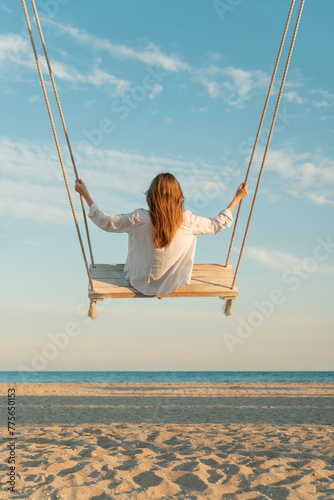 Blond young woman with long hair and white shirt sits on swing above sea. Vertical frame. Yellow magic sunset lights. Back view