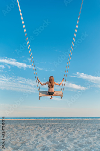 Young woman in bikini swinging at beach over the sea on blue sky background. Vertical frame. Paradise vacation