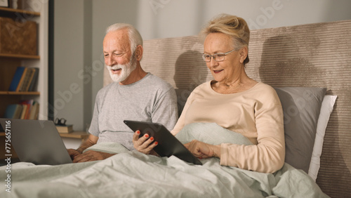 Smiling senior couple using a digital tablet and a laptop in their bed