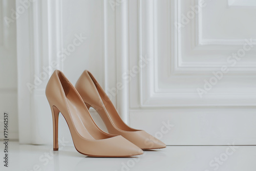 paire de chaussures en cuir de luxe, à talon aiguille, beige, devant un fond représentant un mur blanc avec des moulures en bois, posée sur un sol blanc. Espace négatif copy space photo