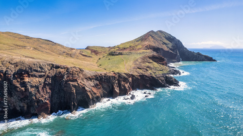 The rocky coast of Madeira, with green cliffs rising above the blue ocean. Waves gently crash against the base of the cliffs, creating white foam. The sky is almost clear with a few clouds.