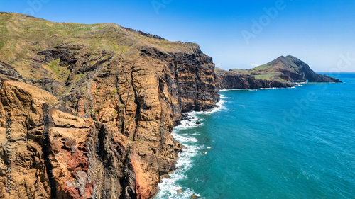 A steep, rocky coastline with light blue ocean water at its base. Visible are green grassy areas and paths along the cliffs. It's the beauty of nature in Madeira, the PR8 trail.
