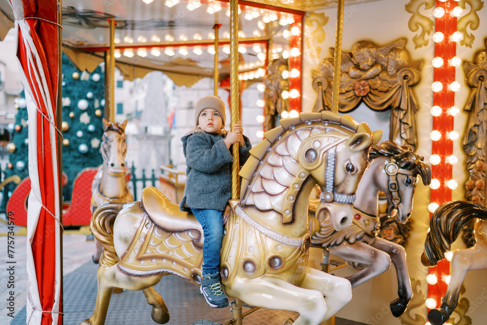 Little girl rides a toy horse on a carousel in the square near a decorated Christmas tree