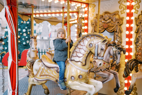 Little girl rides a toy horse on a carousel in the square near a decorated Christmas tree © Nadtochiy