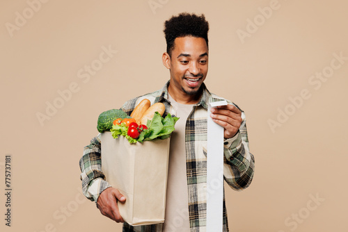Young happy man wear grey shirt hold paper bag for takeaway mock up with food products read cash bill receipt isolated on plain pastel light beige background. Delivery service from shop or restaurant. photo