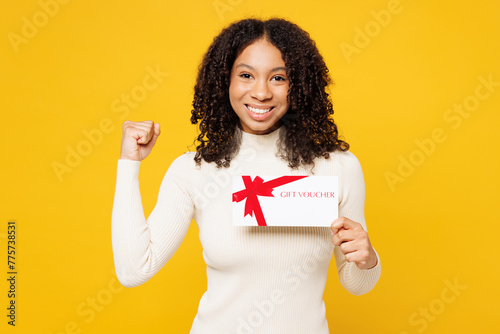 Little kid teen girl of African American ethnicity in white casual clothes hold gift certificate coupon voucher card for store do winner gesture isolated on plain yellow background Childhood concept photo