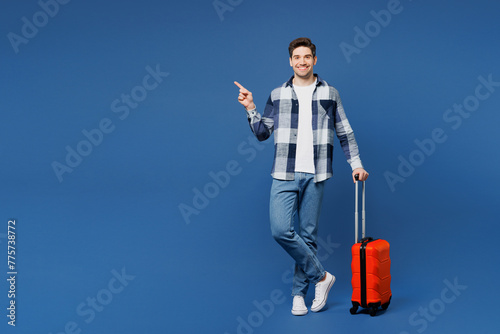 Traveler man wears shirt casual clothes hold suitcase bag point finger aside isolated on plain blue background. Tourist travel abroad in free spare time rest getaway Air flight trip journey concept.