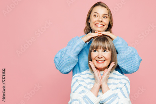 Elder parent mom 50s years old with young adult daughter two women together wear blue casual clothes stand behind mother hold hands on head isolated on plain light pink background. Family day concept. © ViDi Studio
