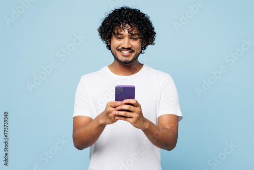 Young fun happy Indian man he wear white t-shirt casual clothes hold in hand use mobile cell phone chat online isolated on plain pastel light blue cyan background studio portrait. Lifestyle concept.