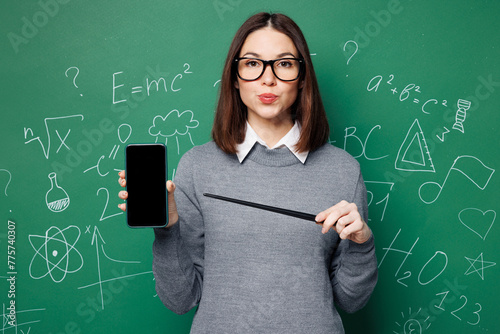 Young smart teacher woman wear grey casual shirt glasses hold use point on blank screen mobile cell phone isolated on green wall chalk blackboard background. Education in high school college concept. © ViDi Studio