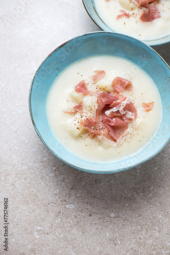 Potato soup with prosciutto in a turquoise bowl, vertical shot with space, elevated view on a beige stone background