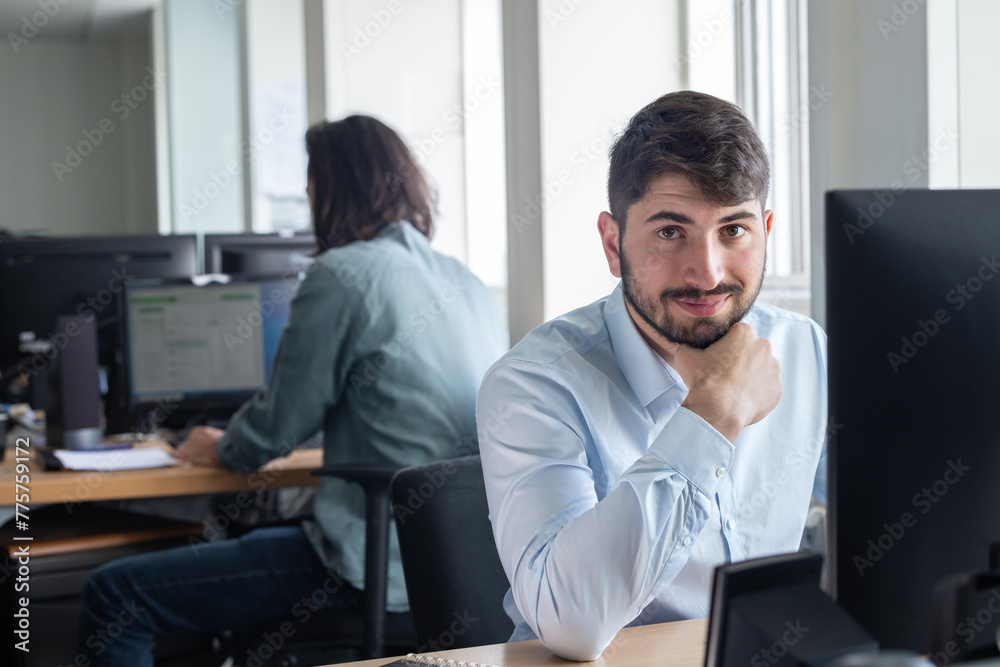 portrait d'un jeune homme employé de bureau souriant quoi est assis à son poste de travail devant son ordinateur. Un collègue est assis derrière lui. portrait d'un jeune homme employé de bureau souriant quoi est assis à son poste de travail devant son ordinateur. Un collègue est assis derrière lui.