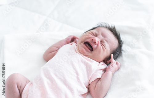 Newborn baby girl crying and screaming, lying on bed at home. Asian Australian infant 19 days old wear clothes, showing unhappy emotion face expression