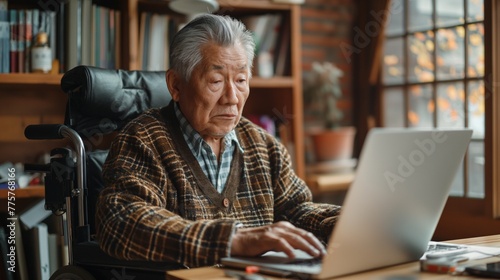 Elderly asian man in a wheelchair at desk working on laptop at home. Disabled person without the ability to move independently. Concept inclusivity at workplace, digital nomad, online education. 
