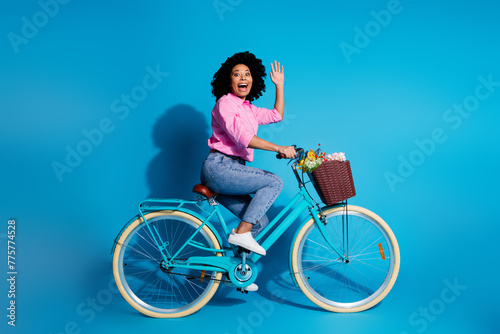 Full length side profile photo of cheerful girl dressed shirt riding bike to empty space waving hand isolated on blue color background © deagreez