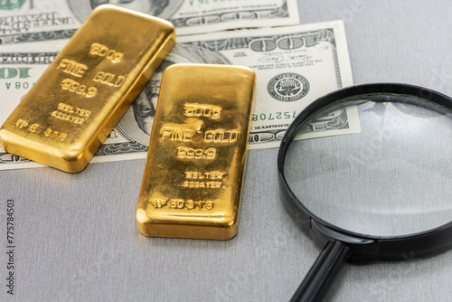 Magnifying glass, gold bars and dollar banknotes on grey desk. Conceptual image of gold trading, buying bullion, checking gold bars