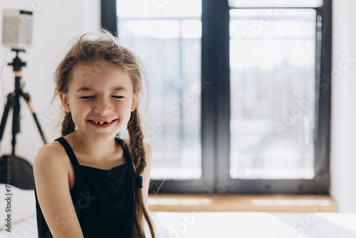 A sweet 6-year-old girl without 1 baby tooth stretches and smiles in the morning in a bright bedroom in front of a large window