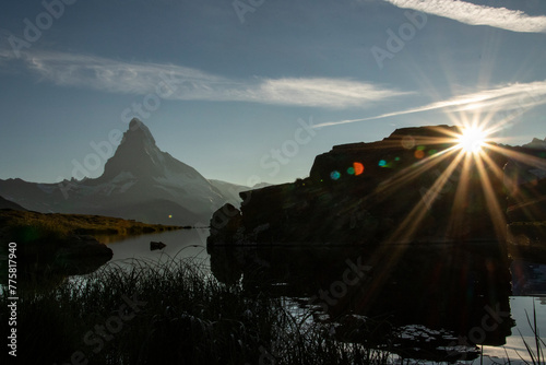 The sun's rays burst over a rocky silhouette, with the iconic Matterhorn reflected in the tranquil waters of Stellisee, heralding a new day in the Swiss Alps photo