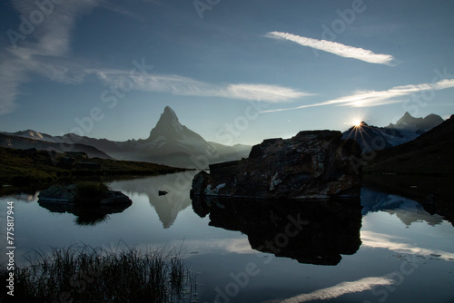 The first rays of sunrise peep over a rugged boulder at Stellisee, with the tranquil waters mirroring the Matterhorn, creating a serene Swiss Alpine scene photo