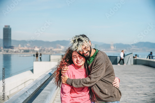 A couple shares a warm embrace, smiling with closed eyes, on a sunny seaside promenade with a soft focus cityscape behind them photo