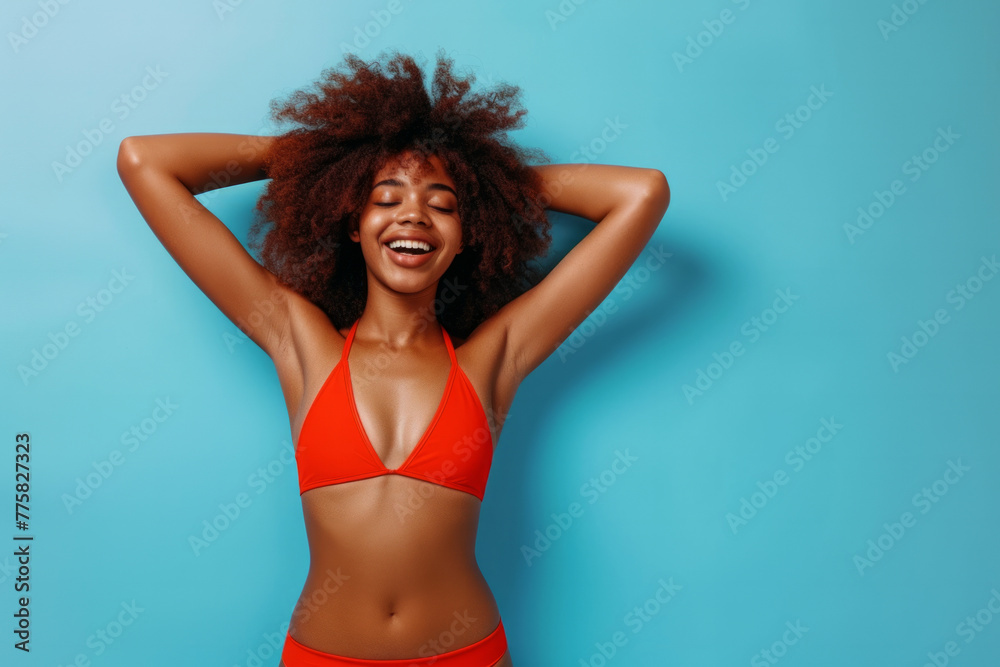 Fototapeta premium Full body portrait of a funny young happy African American woman in red swimsuit going on summer holiday trip and having fun on a blue studio background with copy space. Vacation and travel concept