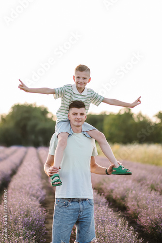 Little son sits on father shoulders, piggyback, in flowers lavender enjoying scent, on summer day at sunset. Child on dad's back on lavender field background. Daddy and boy embrace in meadow.