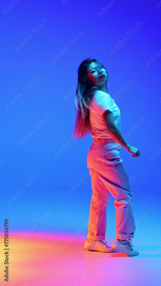 Joyful young Asian woman in casual outfit dancing raising hands against blue gradient background in neon light. Concept of human emotions, self-expression, dance and music, fun and joy. Ad Joyful young Asian woman in casual outfit dancing raising hands against blue gradient background in neon light. Concept of human emotions, self-expression, dance and music, fun and joy. Ad