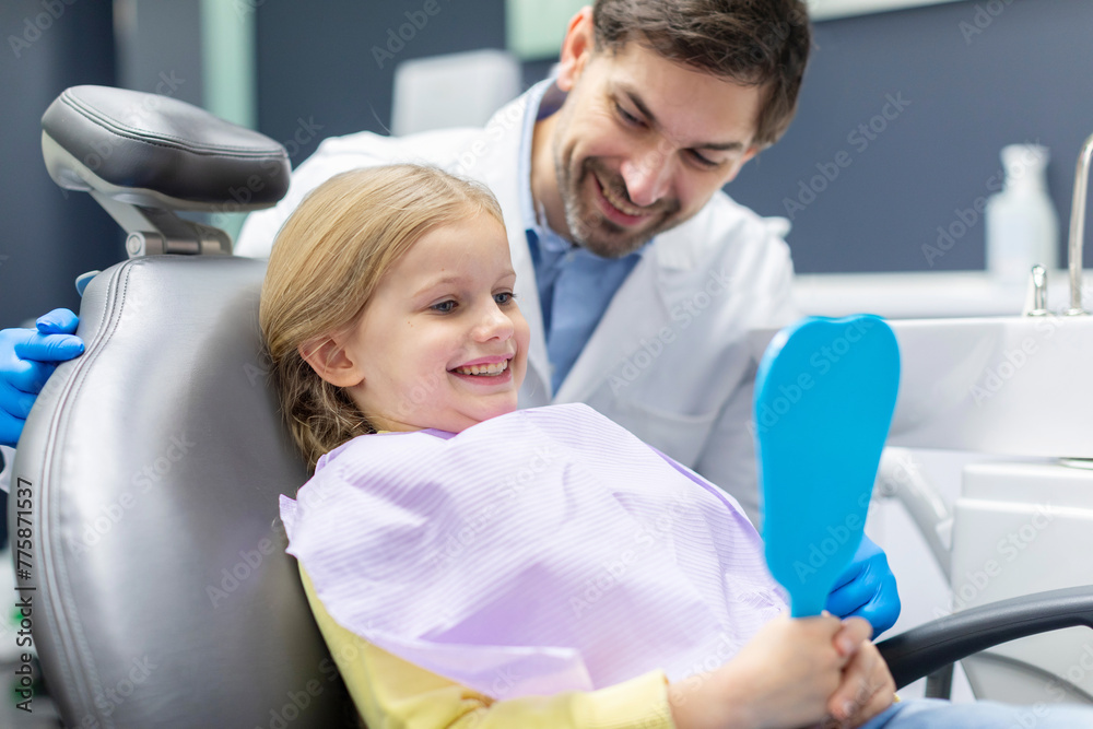Happy little girl in medical chair looking in mirror, male doctor in uniform looks at baby and smiling in modern clinic at daylight Happy little girl in medical chair looking in mirror, male doctor in uniform looks at baby and smiling in modern clinic at daylight