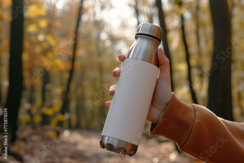 Female hand holding white reusable stainless thermo water bottle in autumn forest.