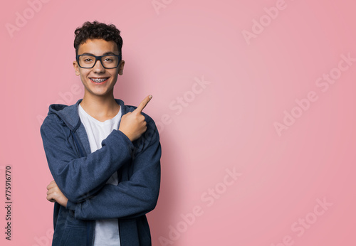 Excited happy black curly haired man in braces brackets, wear glasses, zipup hoodie jaket advertise show point sales slogan text area, isolated rose pink background. Dental care, ophthalmology ad. photo