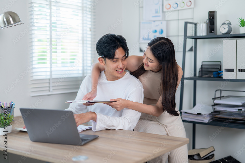 A smiling young Asian couple collaborates on a project, works together managing their budget, working on a laptop in a home office setting. A smiling young Asian couple collaborates on a project, works together managing their budget, working on a laptop in a home office setting.