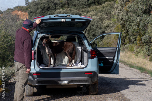 man opening the boot of a 4x4 car where a dog gets in after a walk and looks at him waiting for him to close the door to continue the journey, car on a small mountain road.