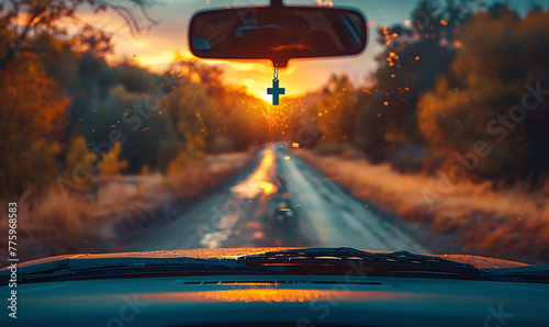 A car’s interior view, driving towards a sunset, cross pendant hanging from the rearview mirror. photo